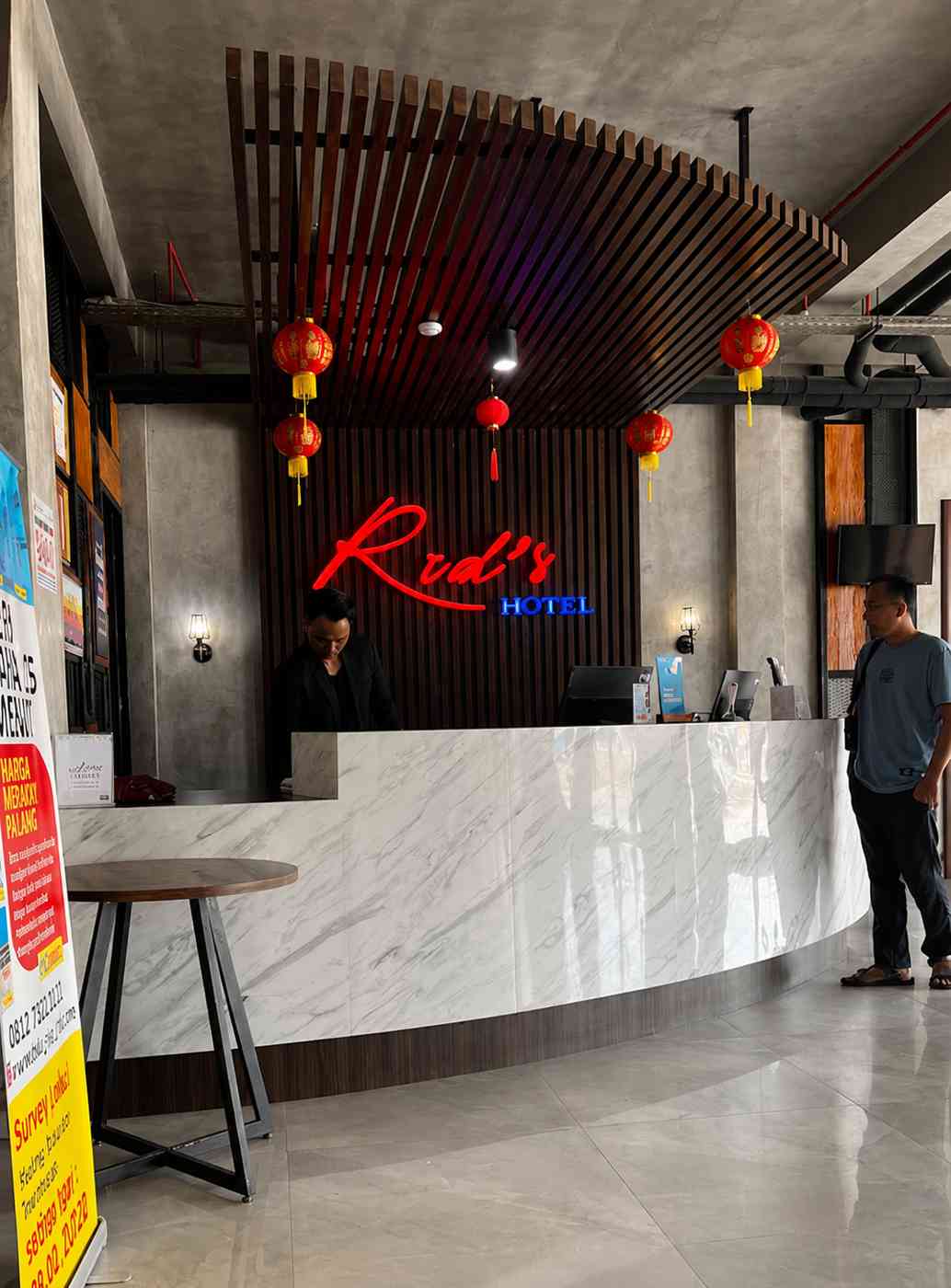 Meja resepsionis bermaterial marmer di bawah papan neon merah Rid's Hotel, dengan staf yang bertugas